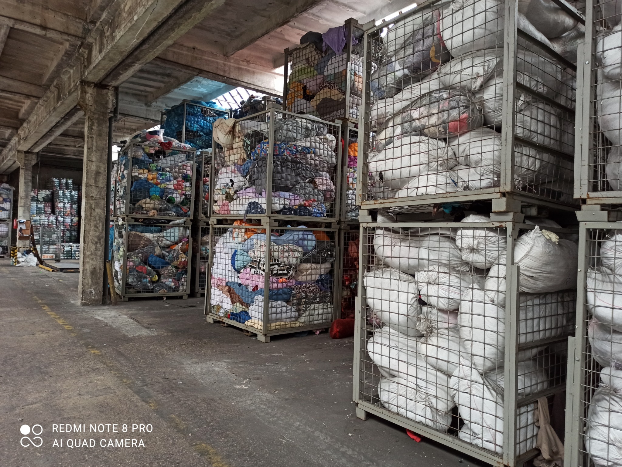 Magazyn z metalowymi koszami wypełnionymi workami i stertami różnorodnej odzieży, widoczne betonowe filary i posadzka.