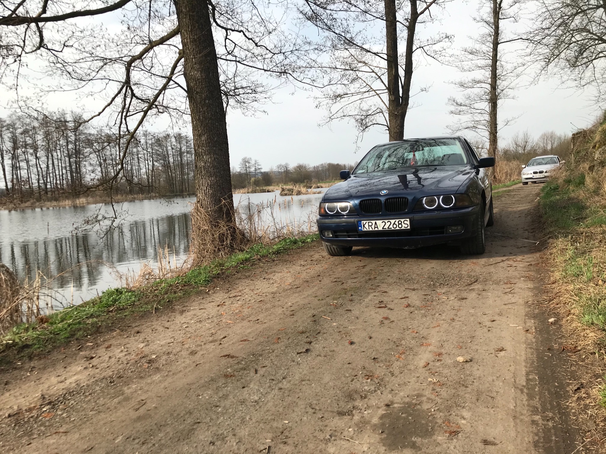 Dwa samochody BMW, ciemnoniebieski i srebrny, zaparkowane na polnej drodze w pobliżu jeziora w pochmurny dzień.