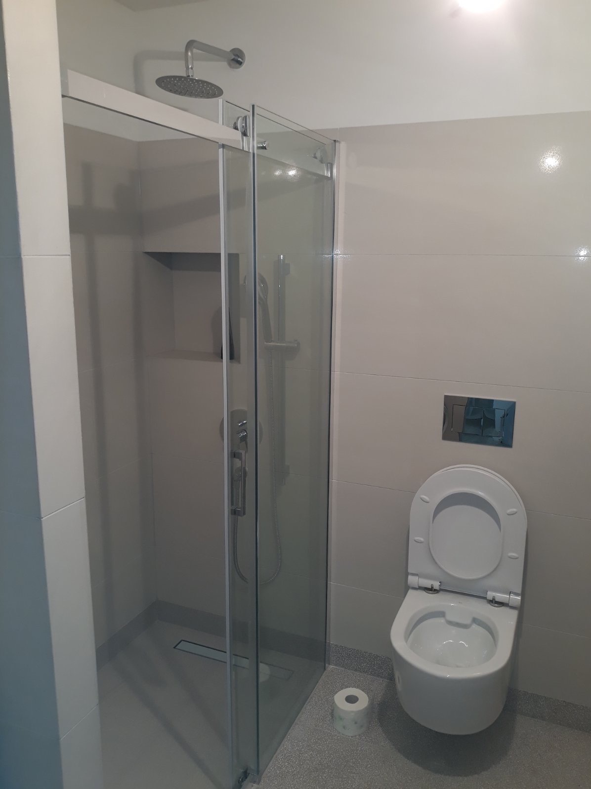 Nowoczesna łazienka z kabiną prysznicową typu walk-in, wiszącą toaletą i rolką papieru toaletowego na podłodze.