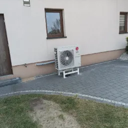 Zewnętrzna jednostka pompy ciepła LG Therma V umieszczona na białym stelażu przy ścianie domu z brązowymi oknami i drzwiami, widoczny szary przewód odprowadzający skropliny, kostka brukowa na podłożu