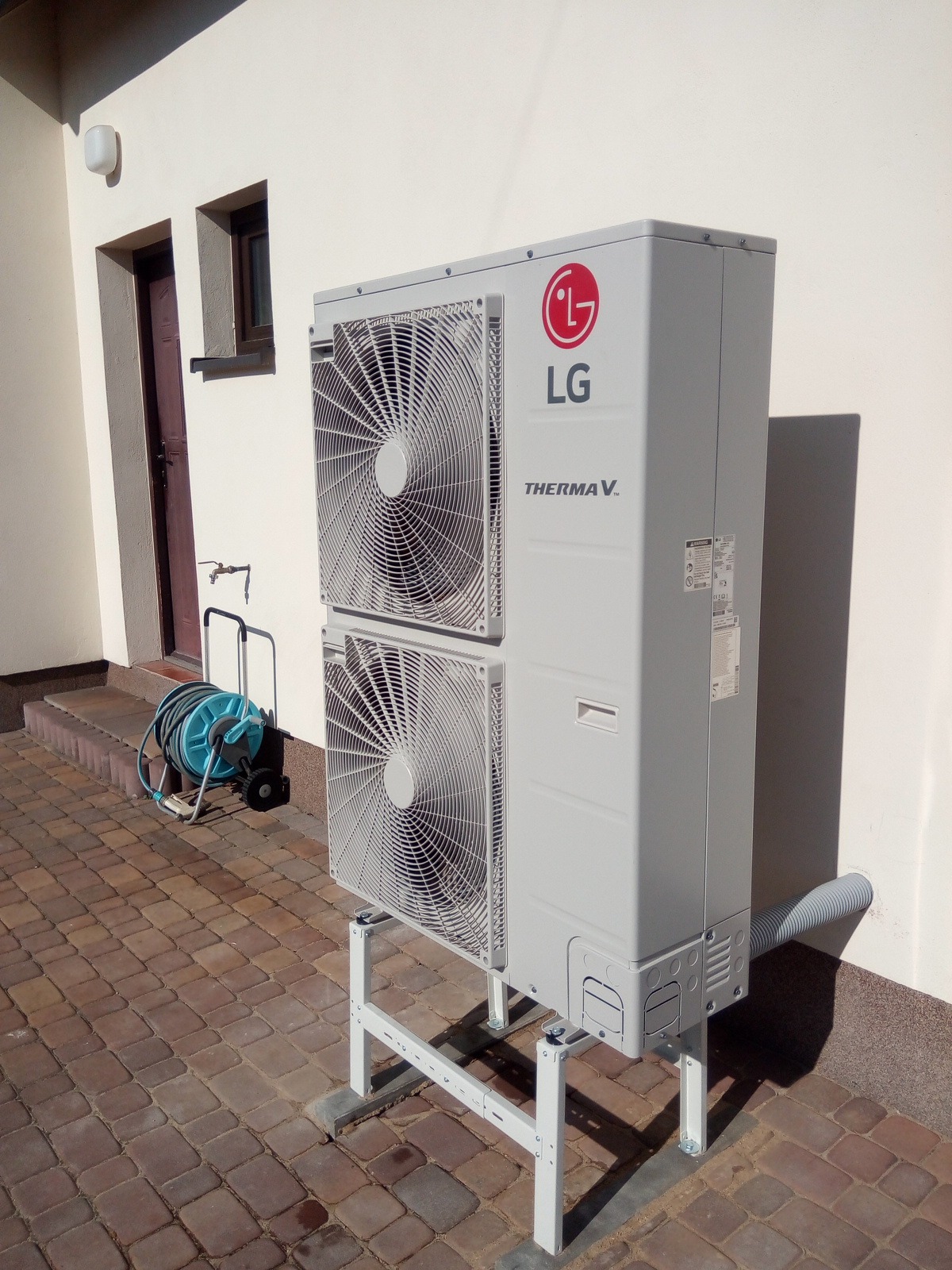 Zewnętrzna jednostka pompy ciepła LG Therma V na metalowym stelażu, umieszczona na kostce brukowej obok budynku, z widocznym wężem ogrodowym na wózku w tle.