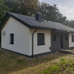 Nowoczesny dom jednorodzinny z białą elewacją i ciemnoszarą stolarką okienną, widoczne palety budowlane przed wejściem.