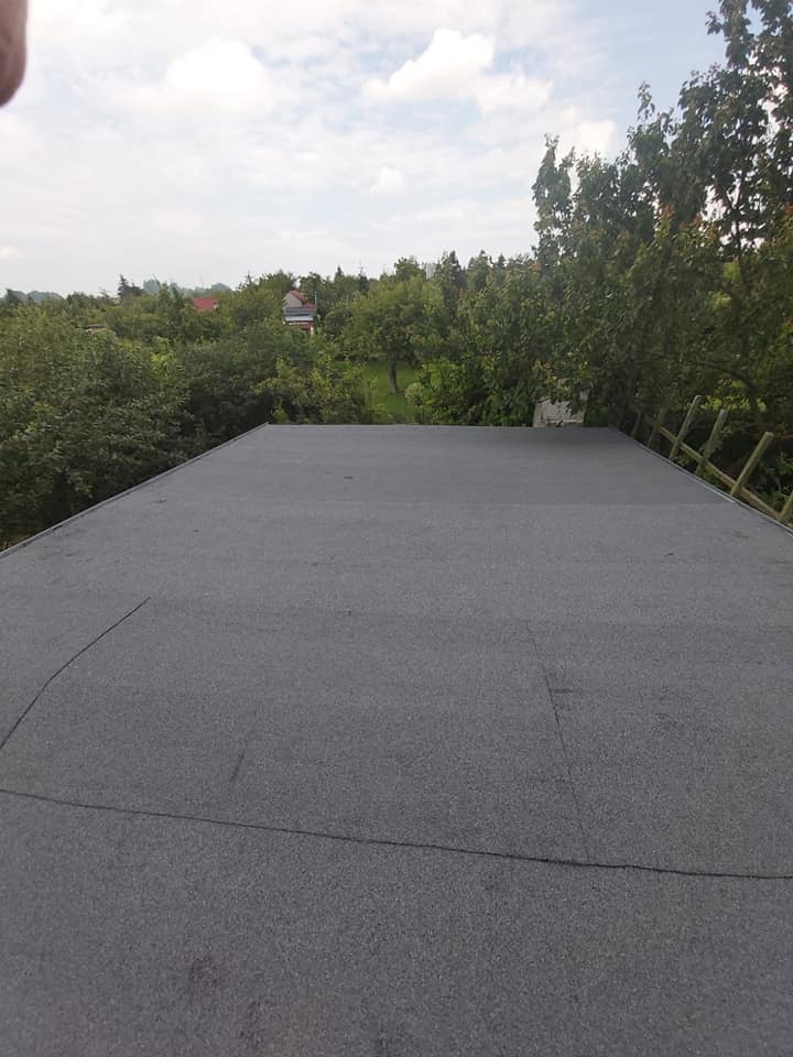 Płaski dach pokryty papą na tle zieleni drzew i błękitnego nieba z chmurami.