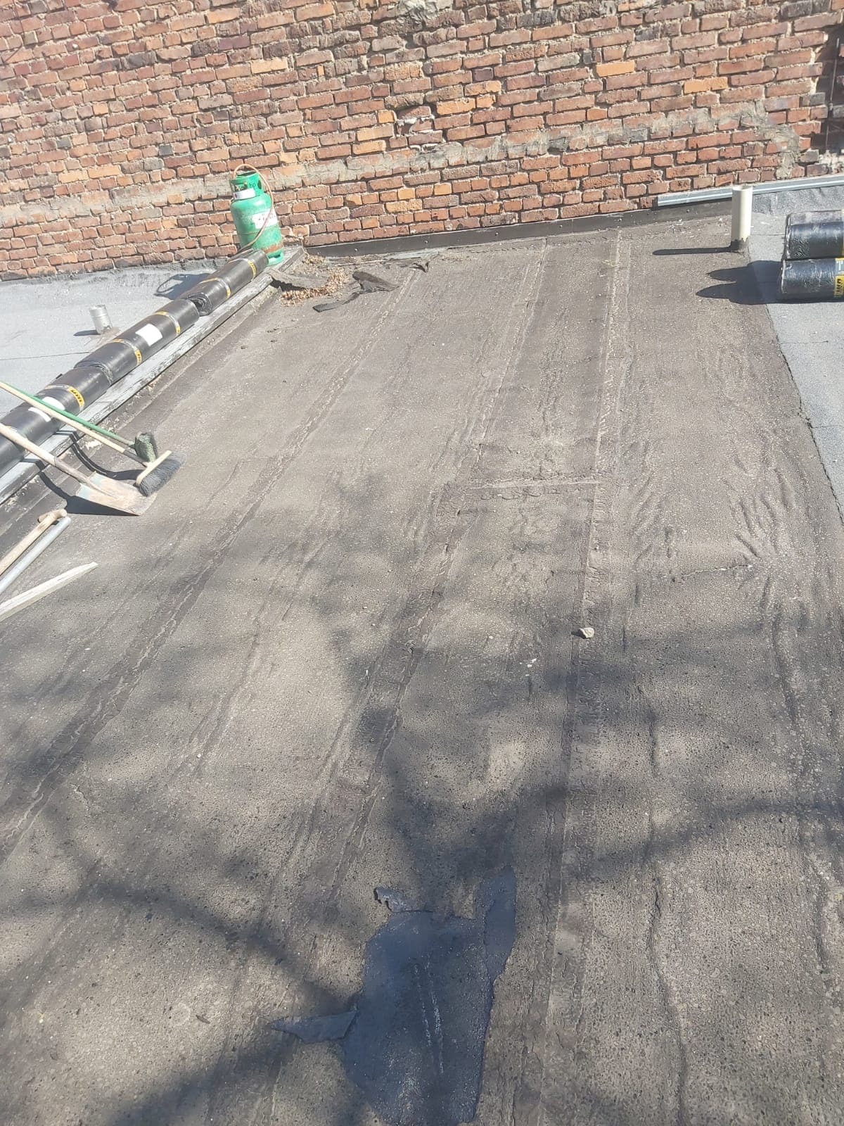 Fragment dachu płaskiego z widocznymi śladami po wałku, butla z gazem, narzędzia dekarskie i rolki papy w tle ceglanej ściany.