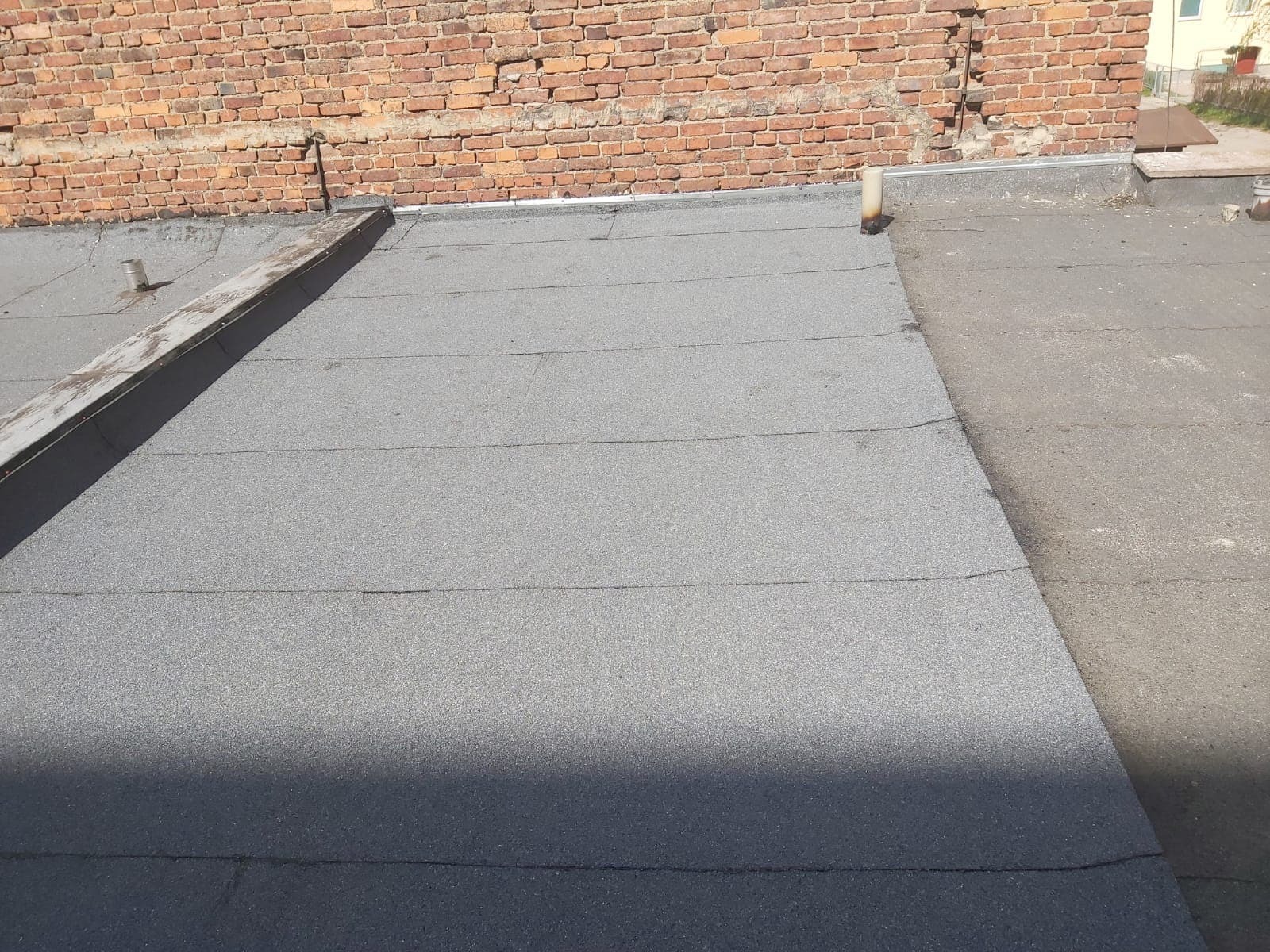 Płaski dach pokryty papą termozgrzewalną, widoczna ceglana ściana budynku i metalowe obróbki blacharskie.