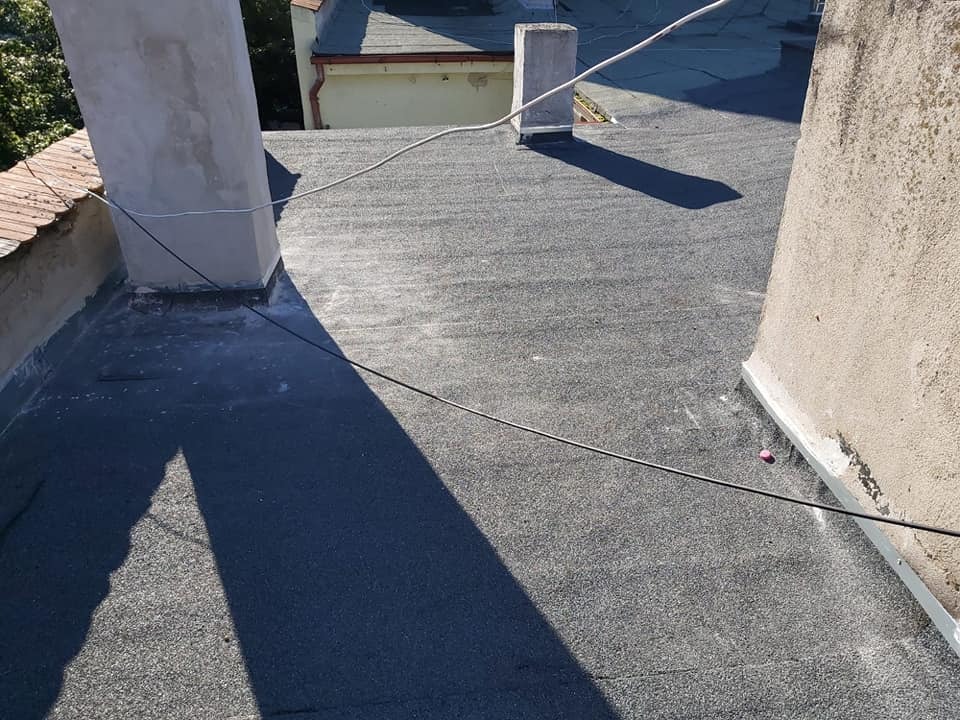 Widok z góry na płaski dach pokryty czarną papą termozgrzewalną, z widocznymi dwoma kominami otynkowanymi na szaro i fragmentem sąsiedniego budynku w tle.