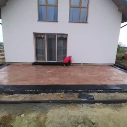 Układanie tarasu z kostki brukowej w kolorze ceglastym przy białym domu z przesuwnymi drzwiami tarasowymi, widoczny pracownik w czerwonej bluzie dopasowujący kostkę