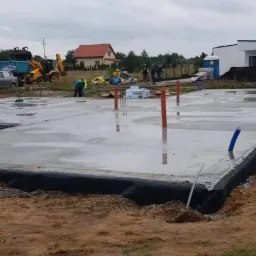 Wylany betonowy fundament z wystającymi rurami instalacyjnymi, zabezpieczony czarną folią, na placu budowy z widocznymi maszynami i ekipą budowlaną w tle.