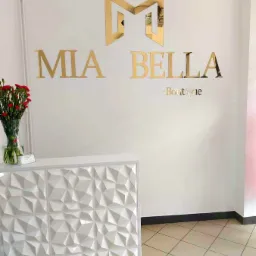 Złote litery z plexi tworzące napis 'MIA BELLA Boutique' i logo w formie litery M, zamontowane na białej ścianie nad białą, trójwymiarową ladą recepcyjną z wazonem czerwonych kwiatów.