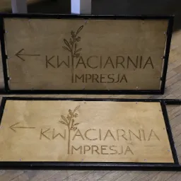 Dwa drewniane szyldy reklamowe z wygrawerowanym napisem 'Kwiaciarnia Impresja' i strzałką, oprawione w czarne metalowe ramy, leżące na drewnianej podłodze.