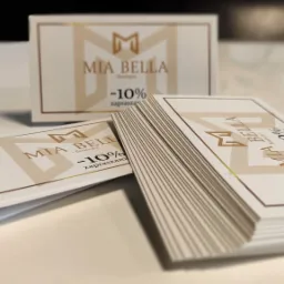 Stos wizytówek rabatowych 'Mia Bella Boutique' z logo i napisem '-10% zapraszam', dodatkowa wizytówka w tle, ułożone na białym, błyszczącym podłożu.