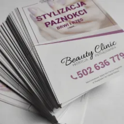 Stos ulotek reklamowych salonu kosmetycznego z ofertą stylizacji paznokci, brwi i rzęs, z widocznym numerem telefonu i adresem.