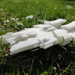Biały, trójwymiarowy model statku kosmicznego, wydrukowany na drukarce 3D, leży na trawie z małymi stokrotkami w słoneczny dzień.