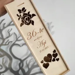 Drewniane pudełko na wino z wygrawerowanym laserowo motywem kwiatowym, sercami i dedykacją urodzinową dla Agi, trzymane w dłoni na tle dekoracji ściennej w kształcie drzewa.