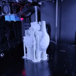 Drukarka 3D w trakcie tworzenia modelu serca z białego filamentu. Widoczne podpory wydruku i dysza drukująca.