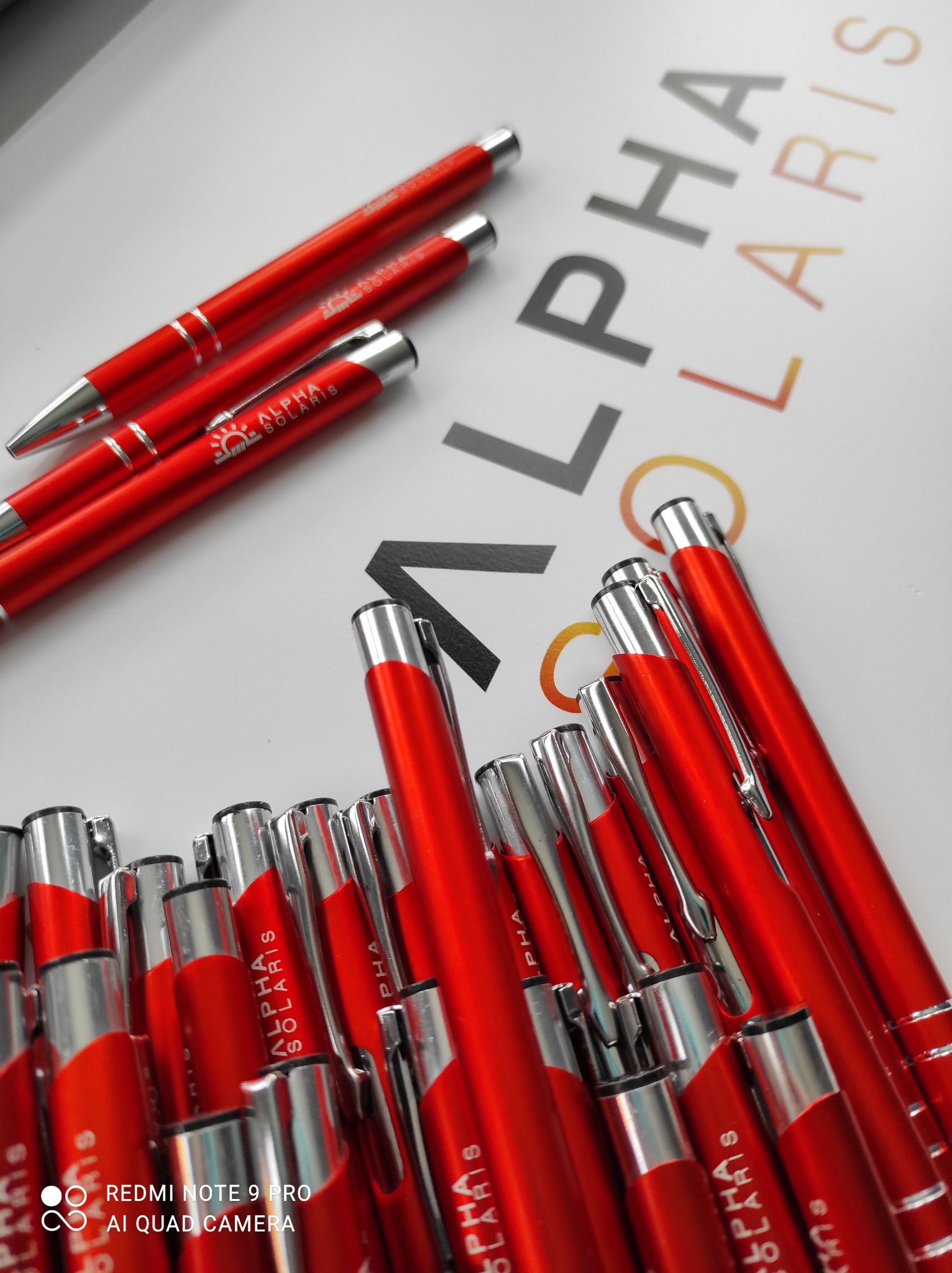 Czerwone długopisy z nadrukowanym logo firmy Alpha Solaris, ułożone na białym tle z częściowo widocznym napisem Alpha Solaris w tle. Długopisy mają srebrne końcówki i klipsy.
