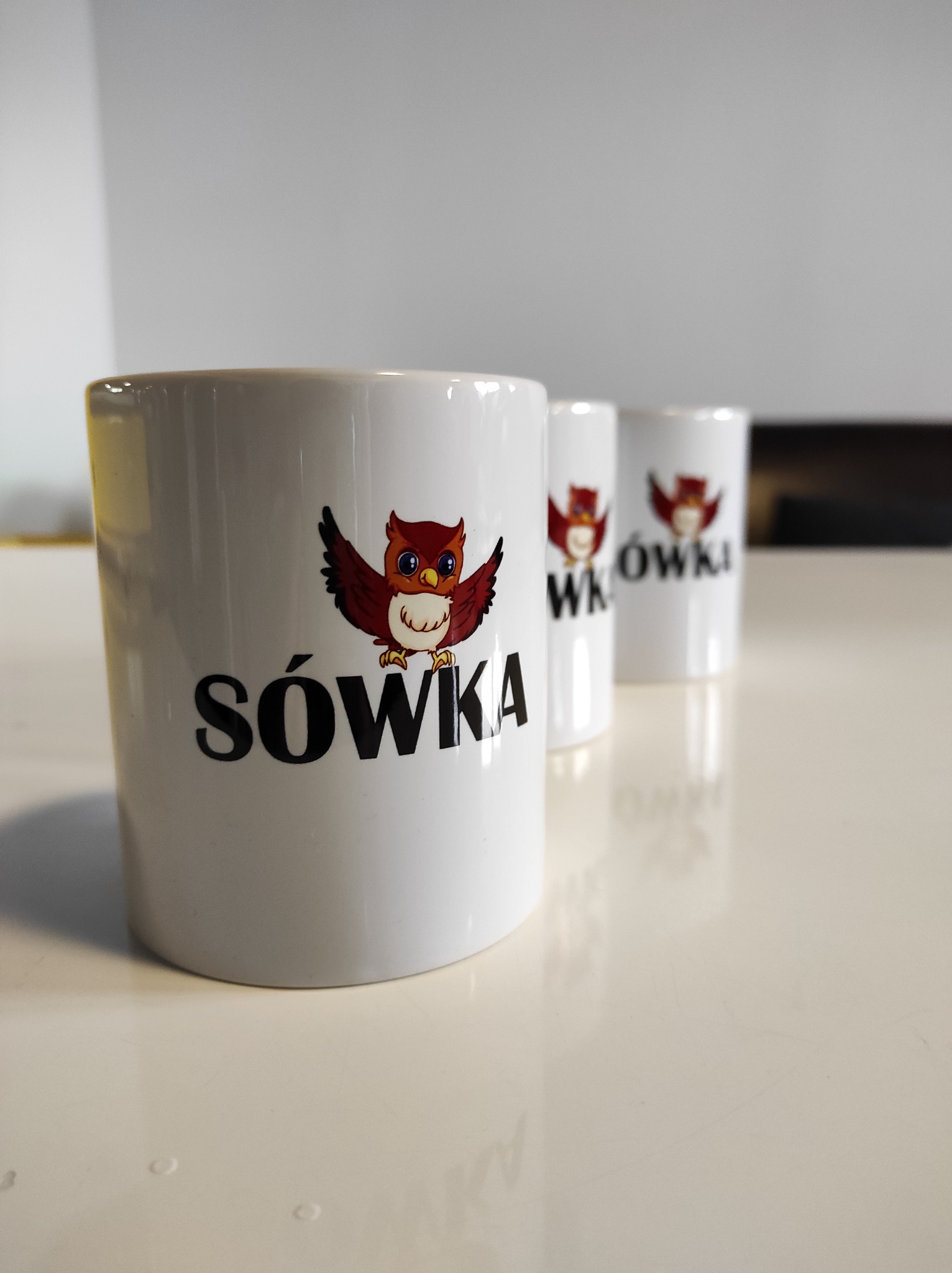 Trzy białe kubki z nadrukowanym logo sowy i napisem 'Sówka', ustawione w rzędzie na jasnym blacie.