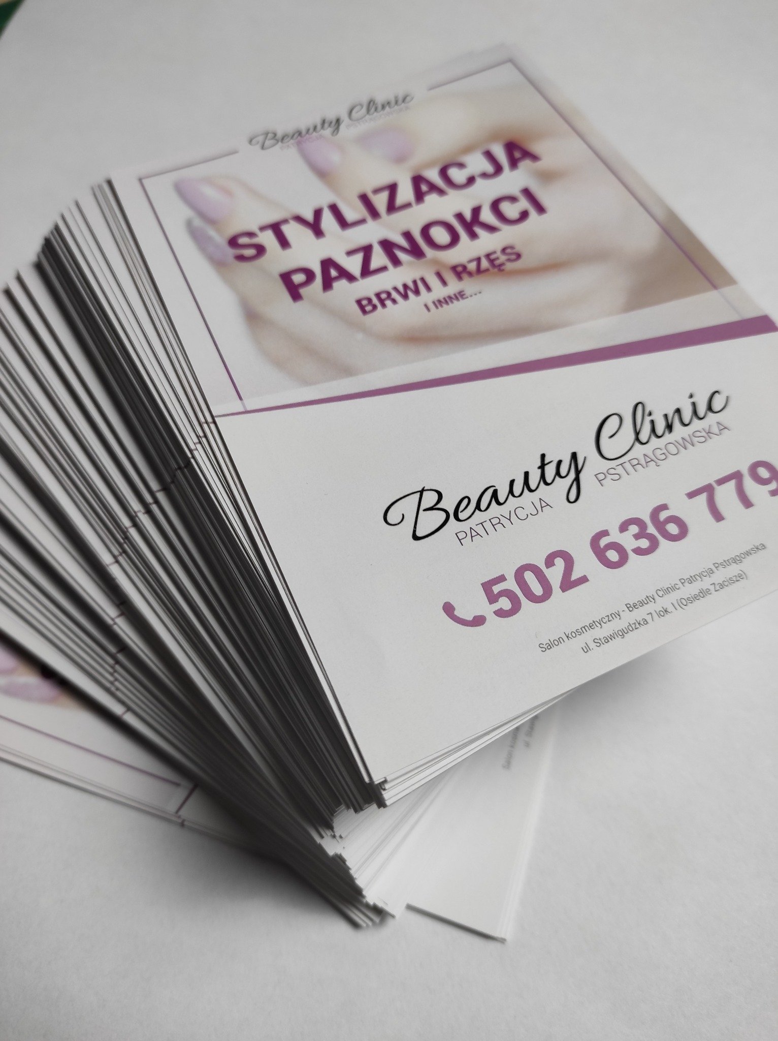 Stos ulotek reklamowych salonu kosmetycznego z ofertą stylizacji paznokci, brwi i rzęs, z widocznym numerem telefonu i adresem.