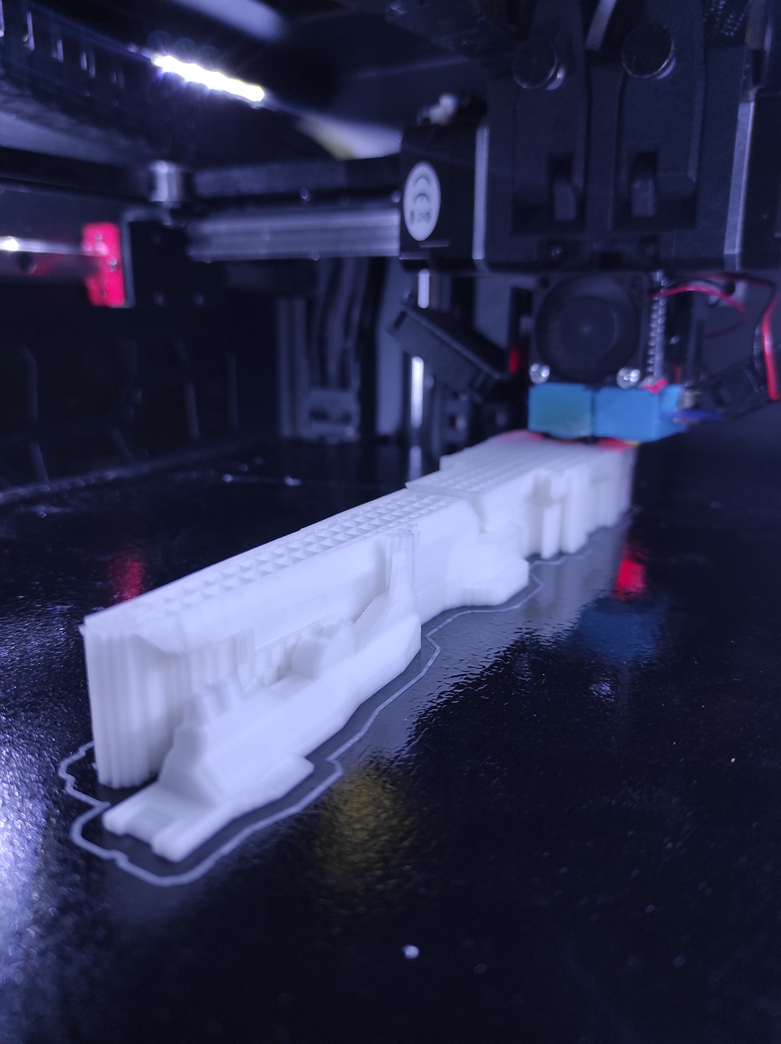 Proces druku 3D: biały, podłużny model z tworzywa sztucznego powstaje warstwa po warstwie na czarnym stole roboczym drukarki 3D, widoczna głowica drukująca i oświetlenie LED.