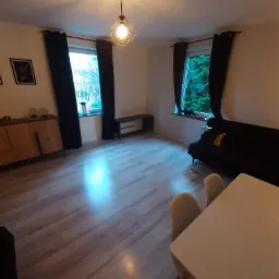 Jasne wnętrze salonu z drewnianą podłogą, czarnymi zasłonami, nowoczesną lampą wiszącą i częściowo widocznym stołem z krzesłami.