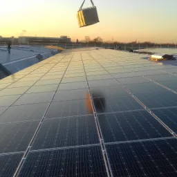 Montaż paneli fotowoltaicznych na dachu budynku przemysłowego; w tle widać budynki, a w górnej części kadru dźwig podnosi kolejny panel.