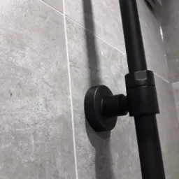 Fragment łazienki z szarymi płytkami i czarnym, metalowym uchwytem zamontowanym na ścianie, widoczne połączenie elementów uchwytu.