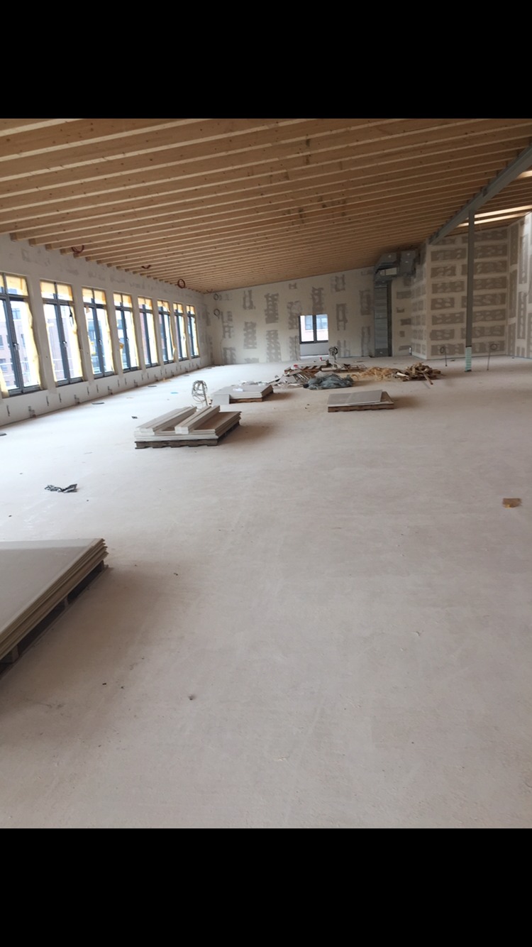 Wnętrze w stanie surowym podczas prac wykończeniowych: widoczne drewniane belki stropowe, okna z folią ochronną, stosy płyt gipsowo-kartonowych na podłodze.