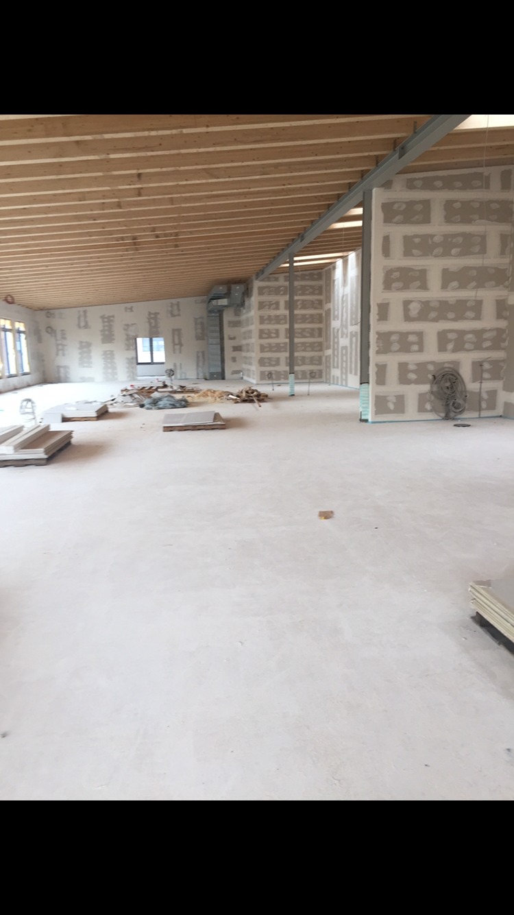 Przestronne wnętrze biurowe w trakcie prac wykończeniowych, widoczne ściany z płyt kartonowo-gipsowych, surowy beton na podłodze, drewniany strop i metalowe elementy konstrukcyjne