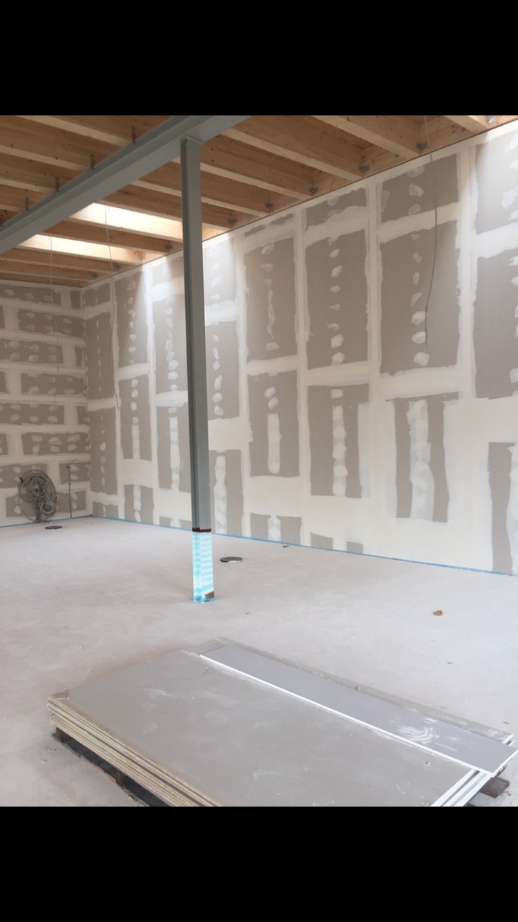 Wnętrze w trakcie prac wykończeniowych: ściany z płyt kartonowo-gipsowych z widocznymi łączeniami, metalowy słup zabezpieczony folią, surowy drewniany strop i stos płyt na podłodze.