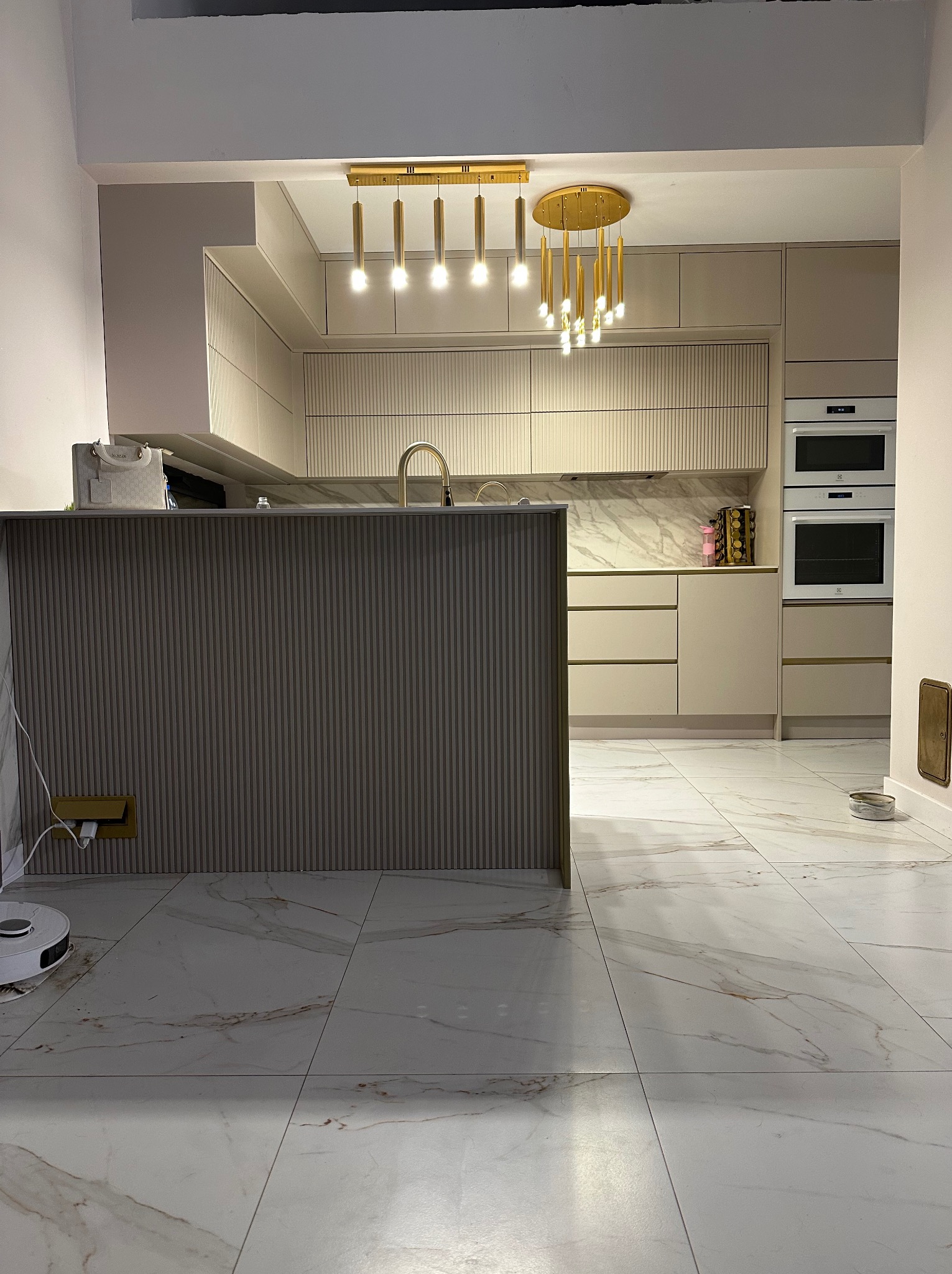 Beżowa kuchnia z ryflowanymi frontami szafek, marmurowym blatem i złotymi detalami oświetlenia, widoczna z perspektywy podłogi z białych płytek z brązowymi żyłami. Dodatkowo widoczny robot...