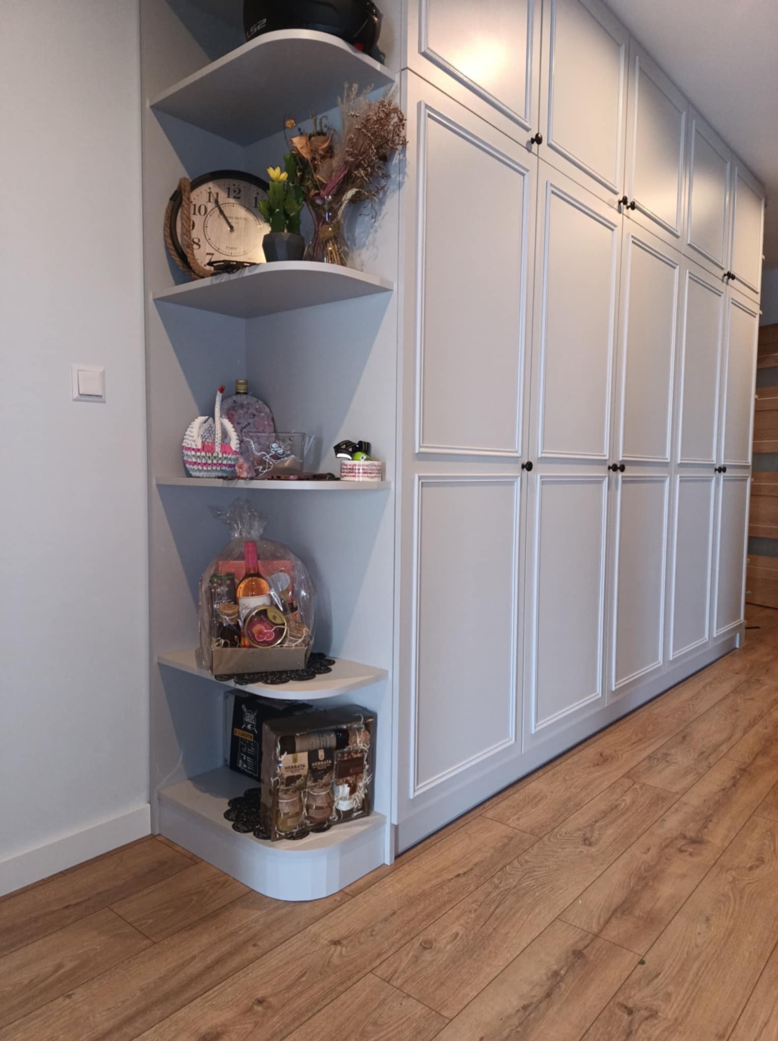 Biała szafa wnękowa z półkami narożnymi, na których stoją dekoracje i prezenty, widok z perspektywy podłogi z paneli.