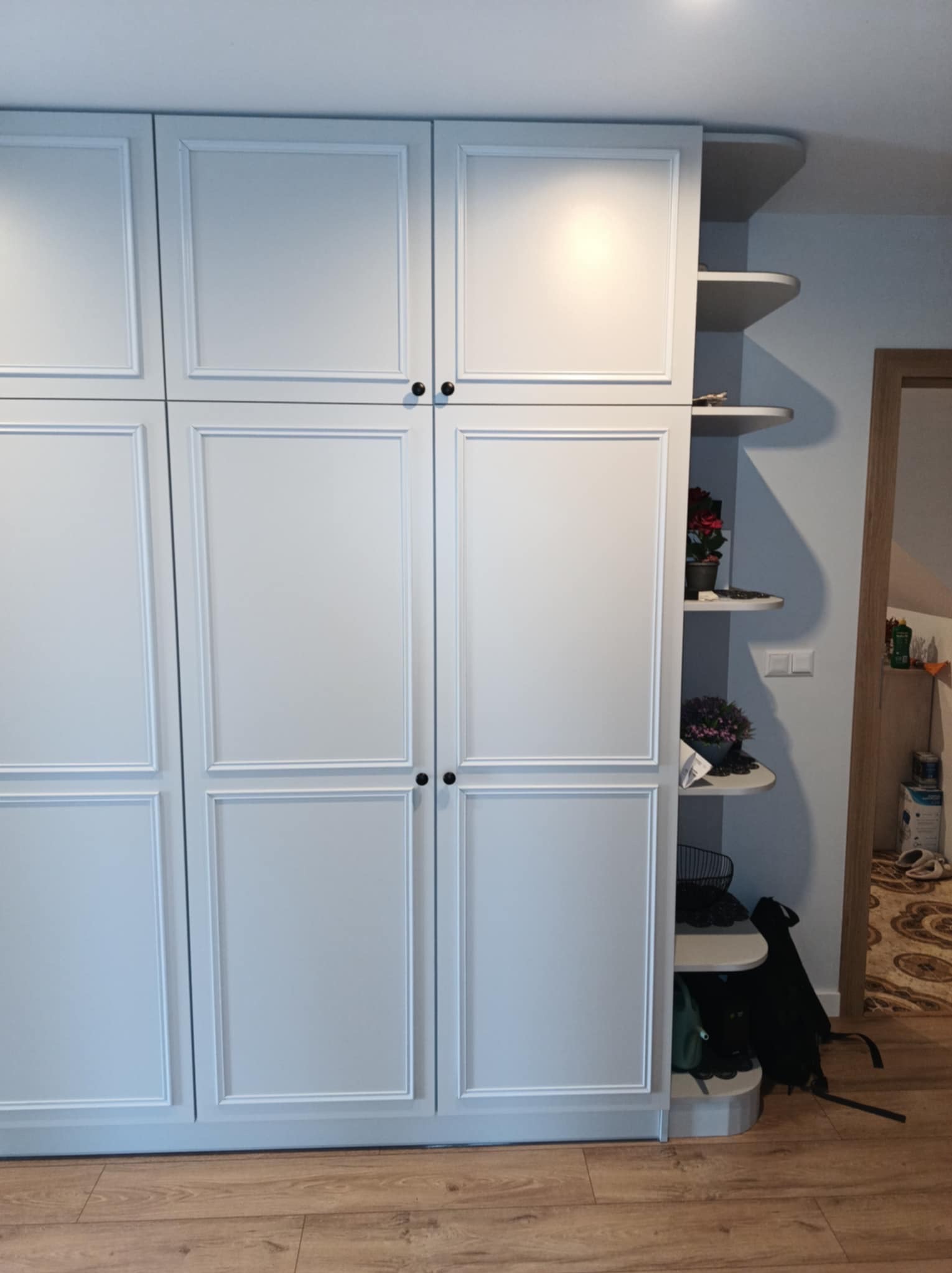 Biała, czterodrzwiowa szafa z frezowanymi frontami i czarnymi gałkami, z otwartymi półkami po prawej stronie, na których stoją doniczki z kwiatami i torba.