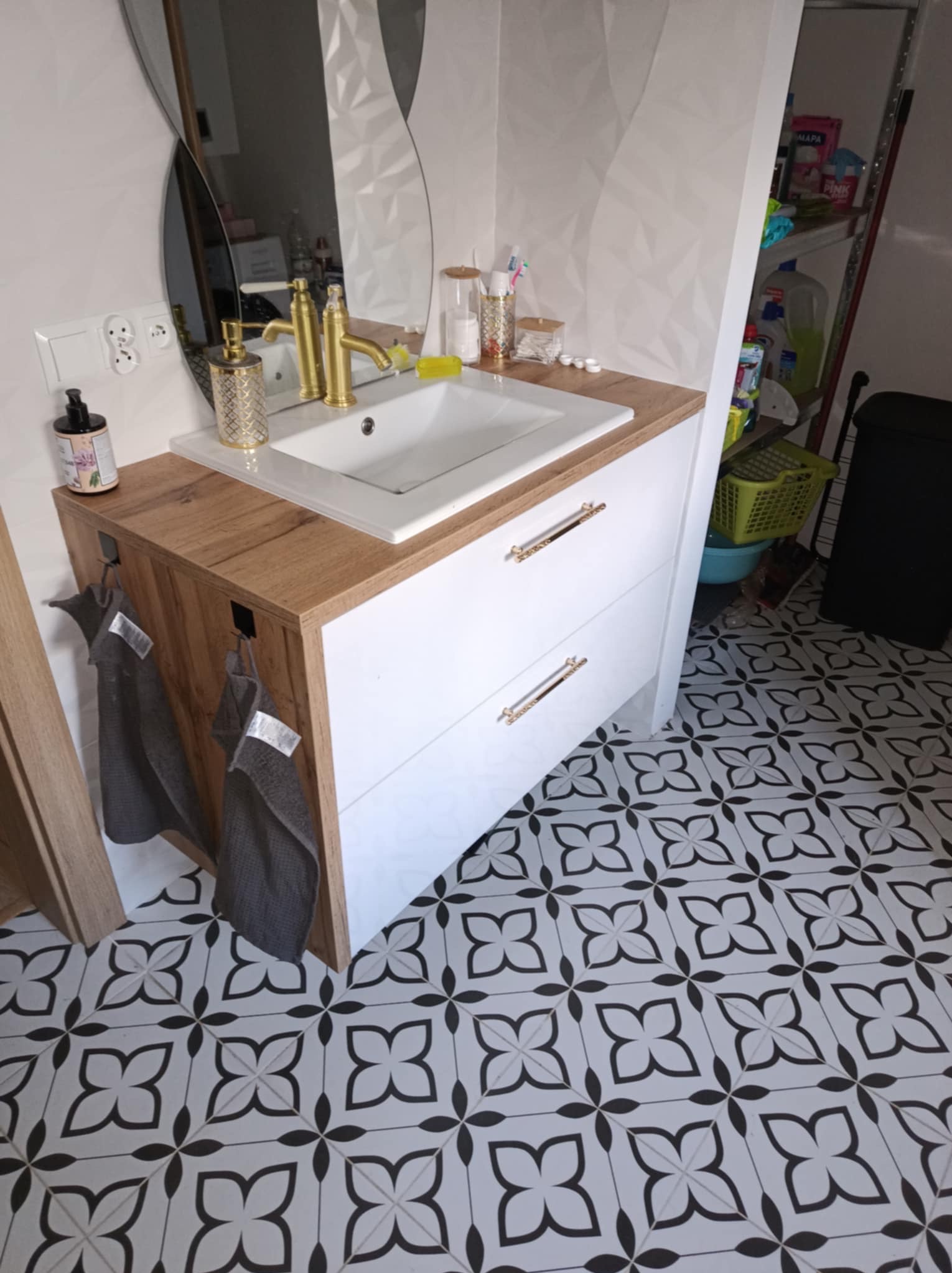 Nowoczesna łazienka z białą szafką pod umywalkę z drewnianym blatem, złotą armaturą i dekoracyjną ścianą w tle, uzupełniona o czarno-białą podłogę w geometryczne wzory.