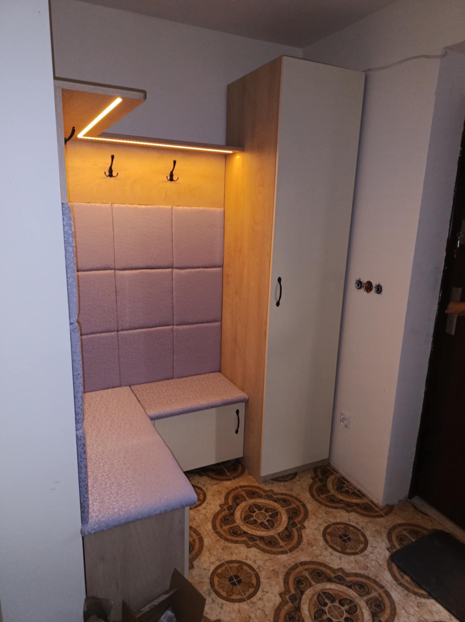 Wnęka w przedpokoju z siedziskiem obitym tapicerką w odcieniach fioletu, oświetleniem LED nad wieszakami i szafą z jasnego drewna, na podłodze brązowe płytki w kwiatowe wzory.