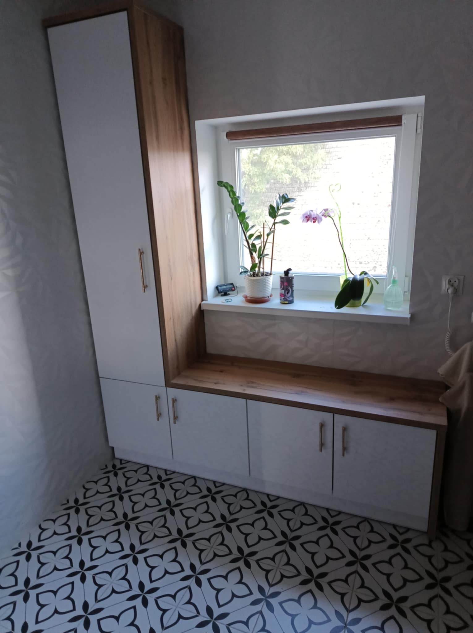 Biało-drewniana szafa z siedziskiem pod oknem, z roślinami doniczkowymi na parapecie, na podłodze wzorzyste płytki.