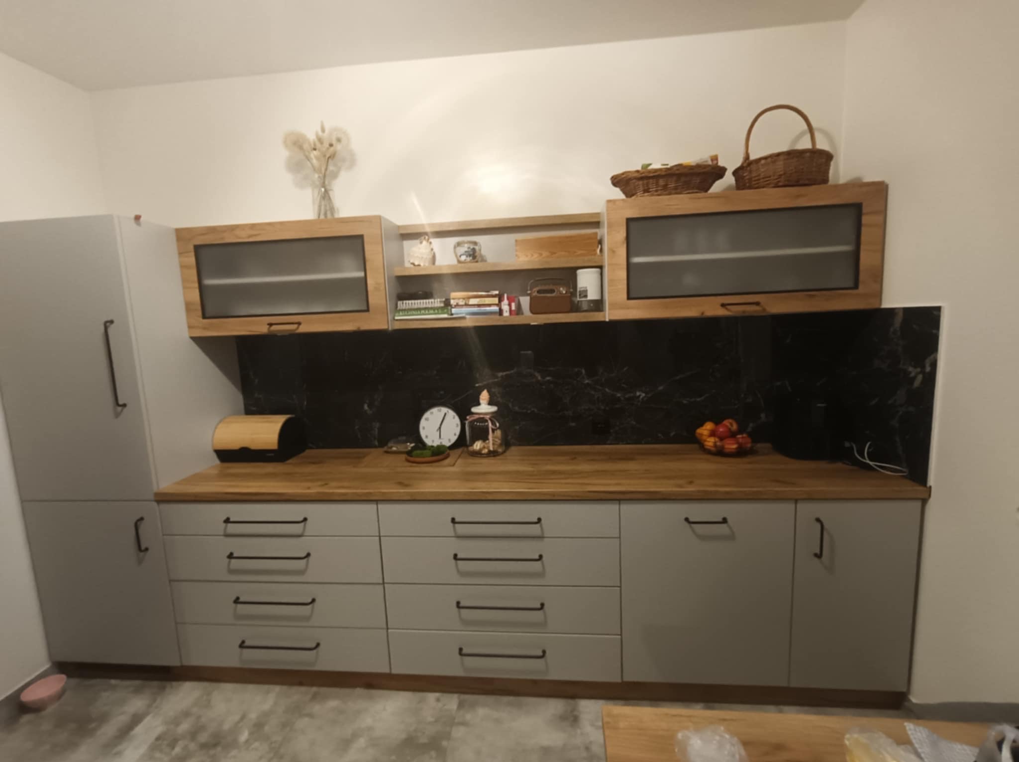 Szare szafki kuchenne z drewnianym blatem i czarnym marmurowym panelem nad blatem, szafki wiszące z przeszklonymi frontami i otwartymi półkami, dekoracje na blacie.