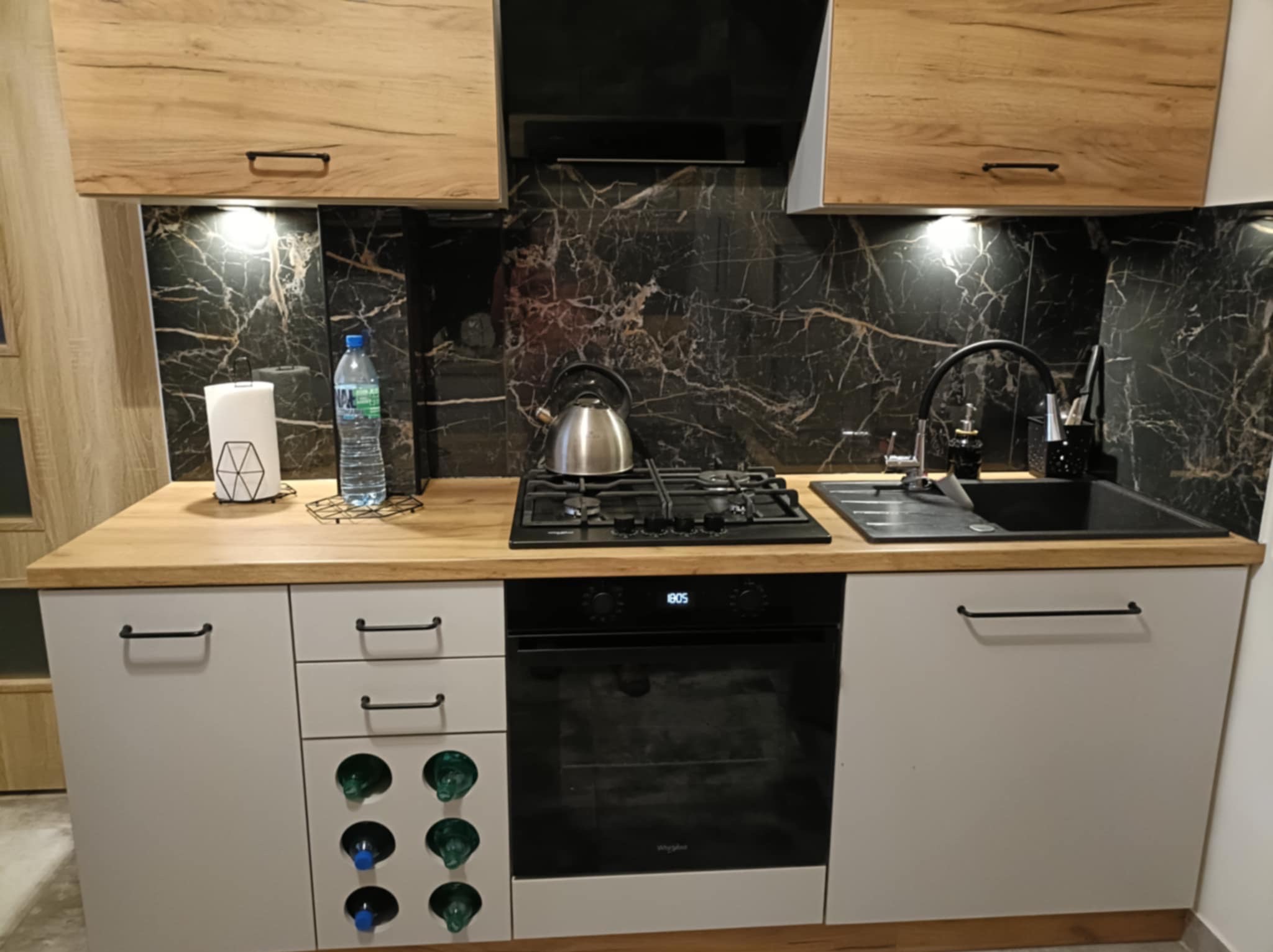 Nowoczesna zabudowa kuchenna w Krakowie z drewnianymi szafkami górnymi, blatem w kolorze drewna i czarnym marmurkowym panelem ściennym. Dolne szafki w kolorze jasnoszarym z czarnymi uchwytami,...