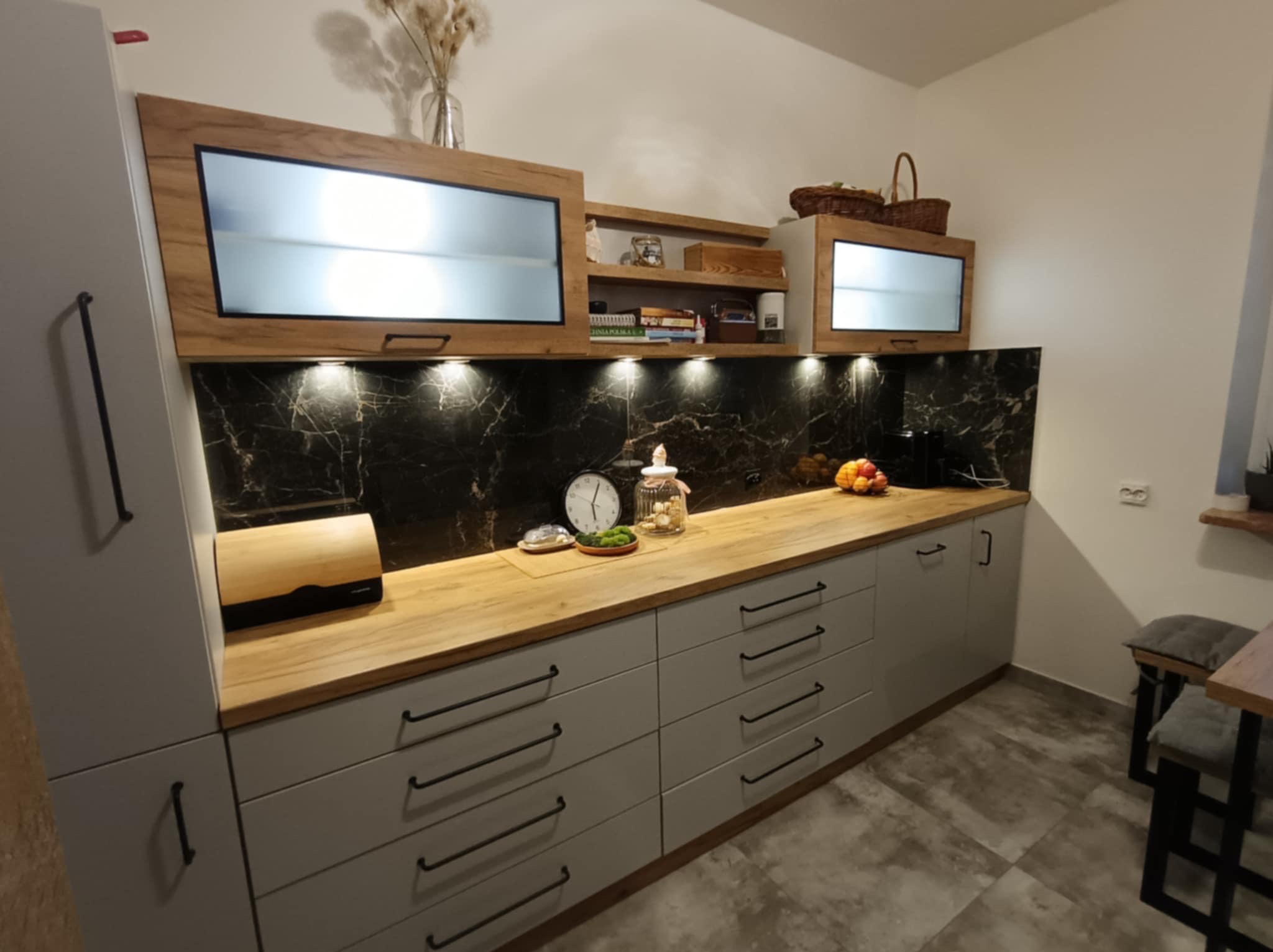 Szara zabudowa kuchenna z drewnianym blatem, czarnym marmurowym panelem i podświetlanymi szafkami wiszącymi. Na blacie chlebak, zegar i ozdobne naczynie.