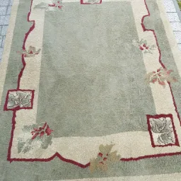 Zielony dywan z kremową bordiurą i motywami kwiatowymi leży na szarej kostce brukowej.