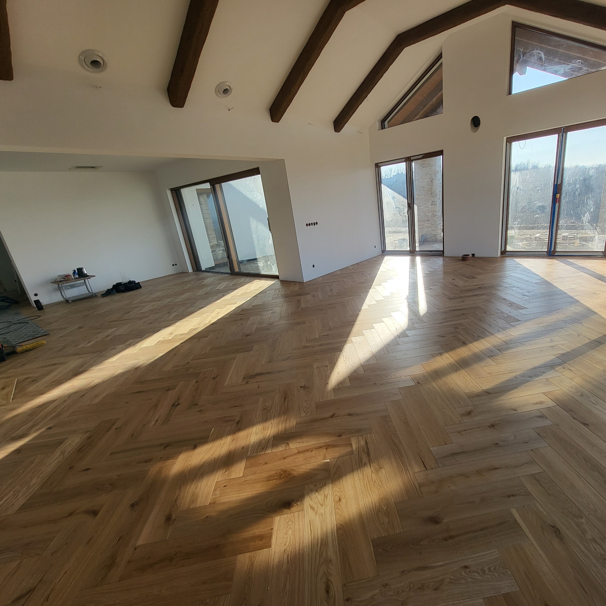 Jasny parkiet ułożony we wzór jodełki francuskiej w przestronnym, jasnym salonie z dużymi oknami i drewnianymi belkami stropowymi.