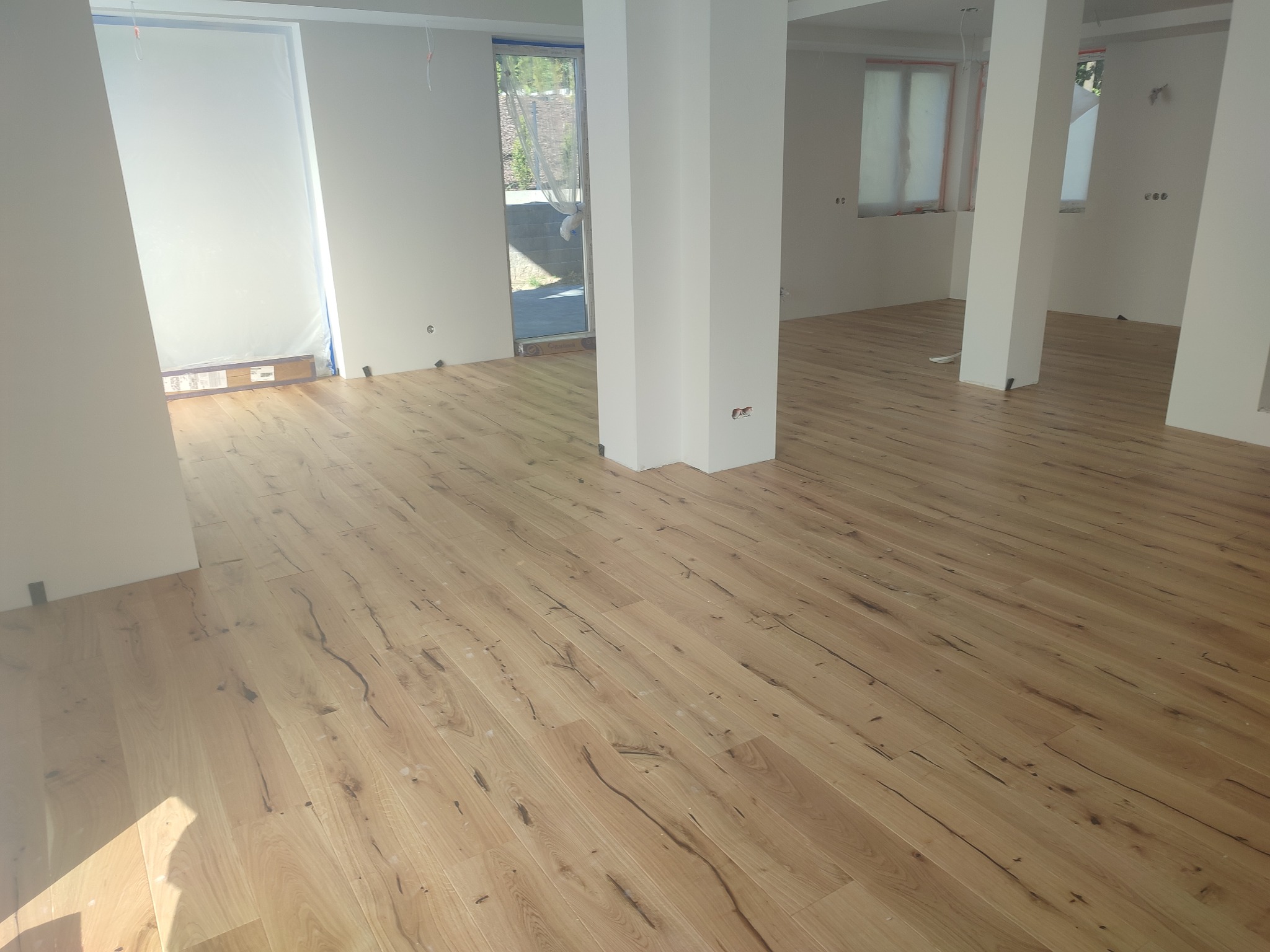 Jasne, drewniane panele podłogowe ułożone w pustym, odświeżonym pokoju z białymi ścianami i kolumnami, okna zabezpieczone folią malarską.