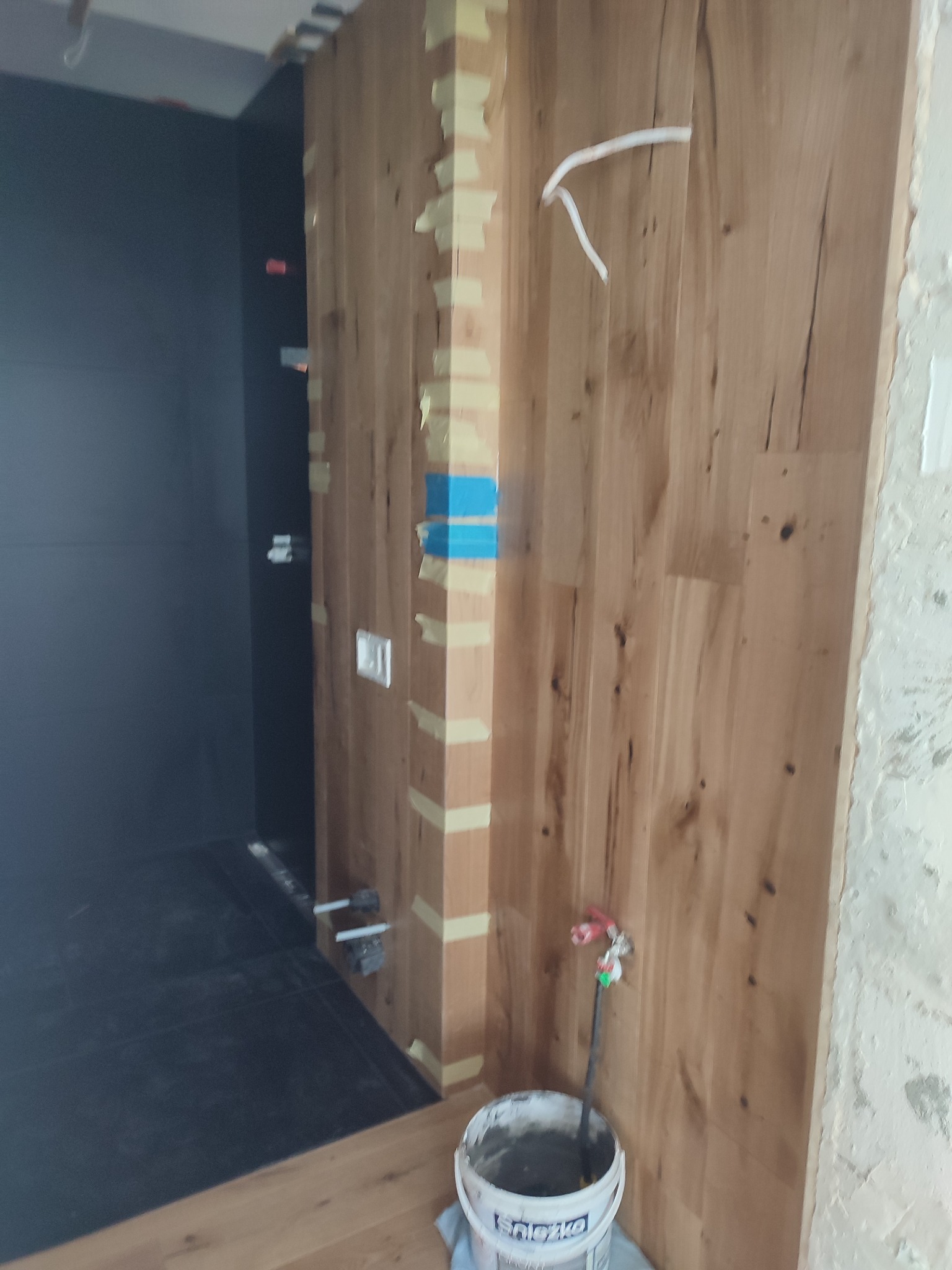 Wnętrze w trakcie prac wykończeniowych: drewniana okładzina ściany zabezpieczona taśmą malarską, widoczne elementy instalacji, wiadro z zaprawą.