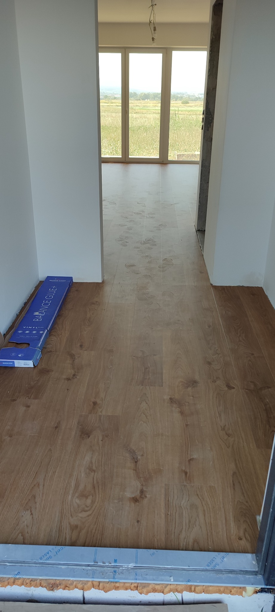 Nowo położona podłoga z paneli winylowych o wzorze drewna, widoczne otwarte opakowanie paneli przy ścianie, ślady stóp na podłodze w tle, widok na łąkę przez okno.