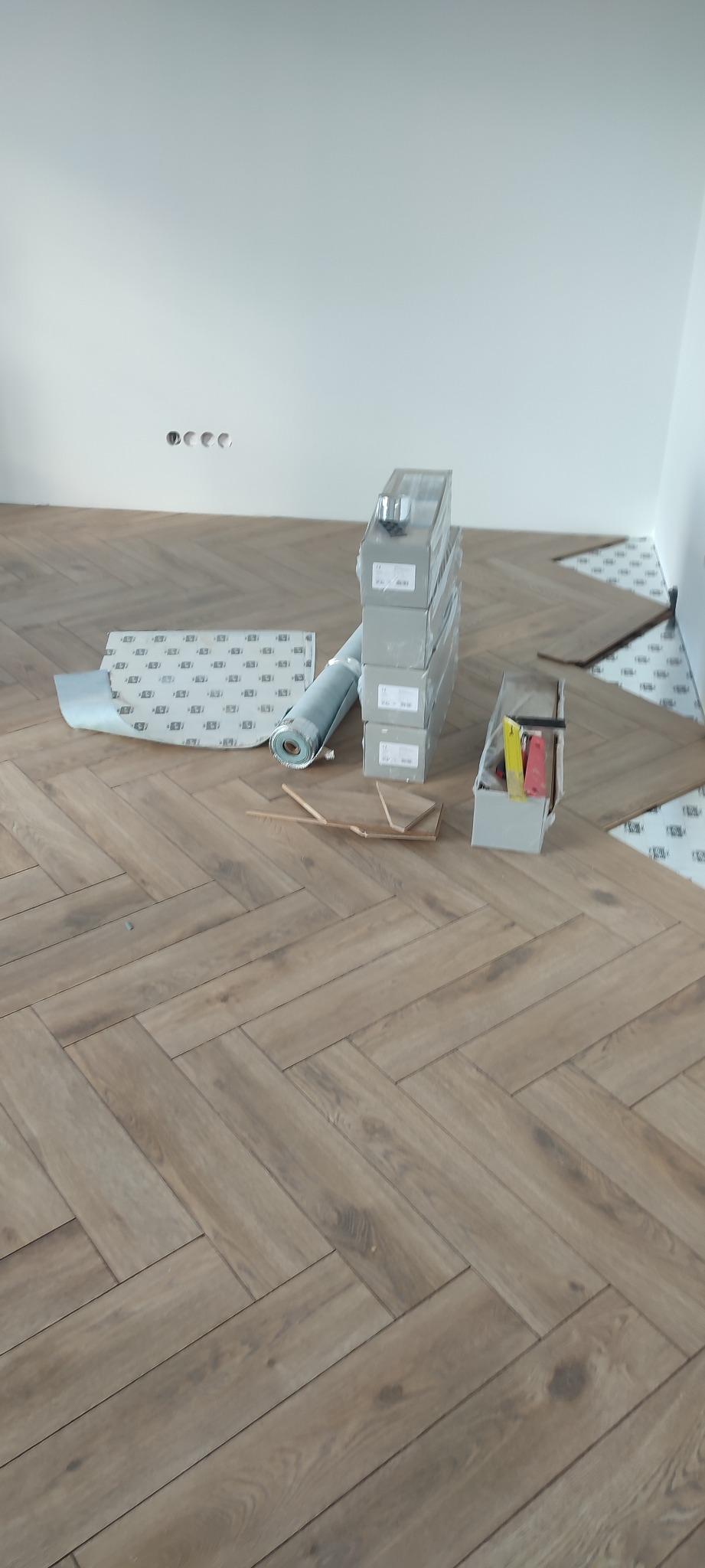 Fragment podłogi z ułożonym parkietem w jodełkę, widoczne materiały przygotowane do dalszej pracy, w tym rolka podkładu i paczki parkietu, oraz narzędzia w pojemniku.