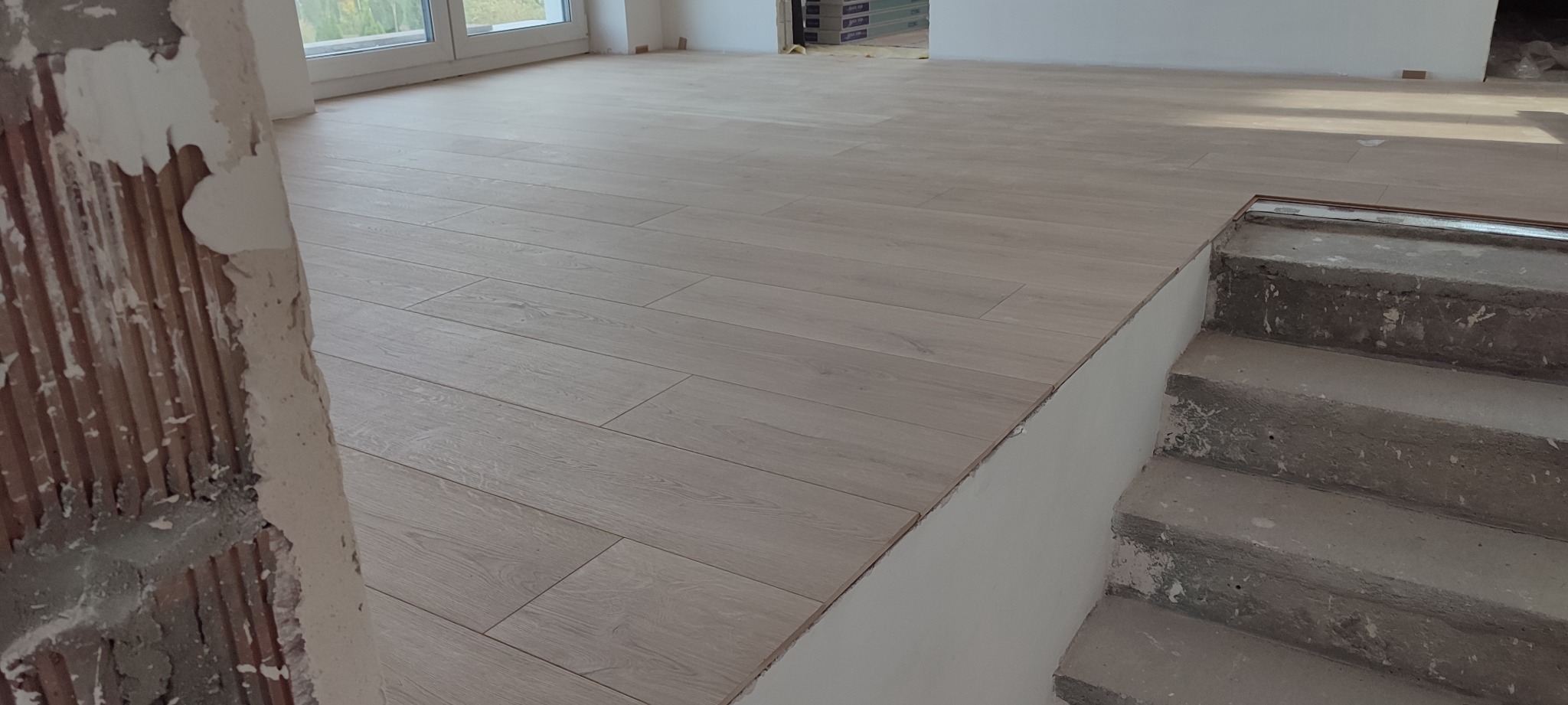 Jasne panele podłogowe ułożone w nowym pomieszczeniu, widoczne surowe schody betonowe i fragment ściany w trakcie wykończenia.