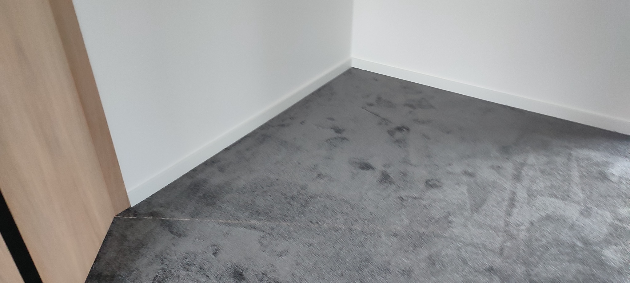 Fragment wnętrza z wykończoną podłogą o szarym, melanżowym wzorze, widoczny fragment drzwi z okleiną drewnopodobną oraz biała listwa przypodłogowa przy ścianie.