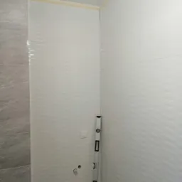 Świeżo ułożone, białe, faliste płytki ścienne w rogu łazienki, z widoczną poziomicą opartą o ścianę, przygotowane podłączenia hydrauliczne i fragment szarej ściany z płytek imitujących marmur.