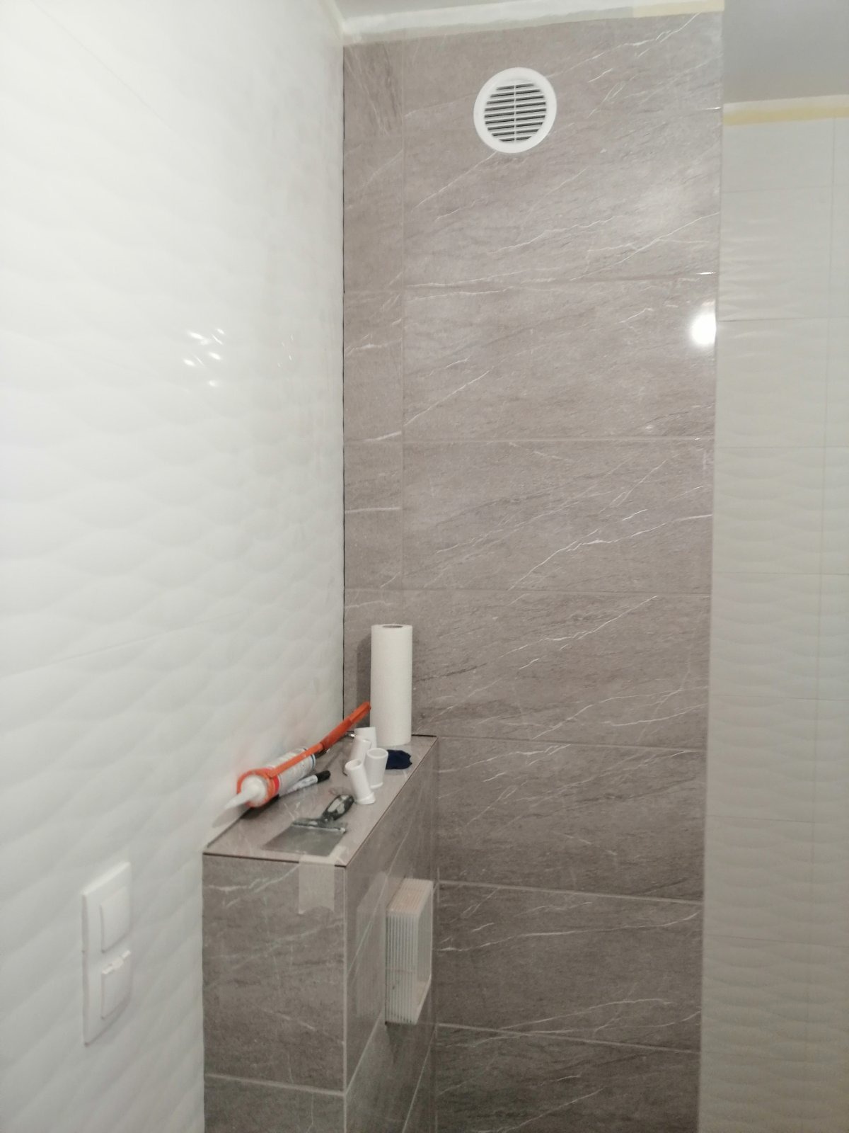 Fragment łazienki w trakcie remontu: szara, marmurkowa glazura ścienna, biała ściana z falistą fakturą, narzędzia i materiały budowlane na obudowie stelaża podtynkowego WC.