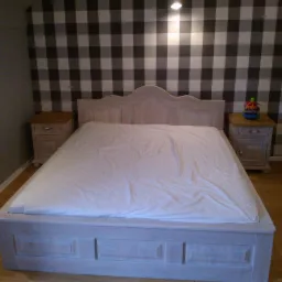 Drewniane łóżko z białym prześcieradłem i szafkami nocnymi na tle ściany w kratę.