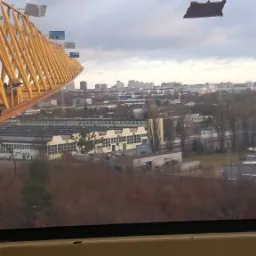 Widok z kabiny żółtego żurawia budowlanego na panoramę miasta z fabrykami i blokami w pochmurny dzień, z widocznymi zabrudzeniami na szybie kabiny.